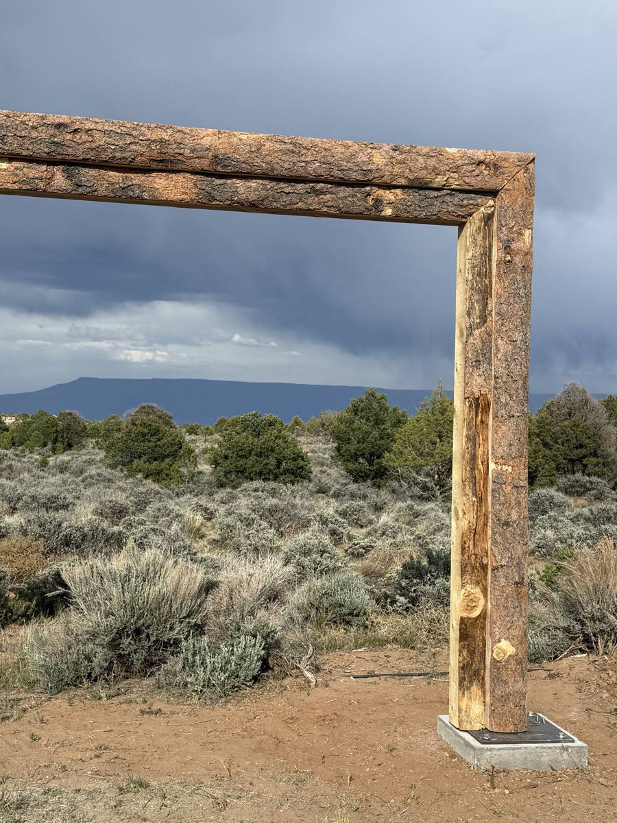 A natural-edge timber frame on a steel base plate, with sagebrush and a flat-topped mesa behind